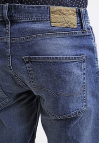 Blå denimjeans med en texturerad brun läderlapp vid midjan, distinkta sömmar på bakfickorna och en klassisk passform.