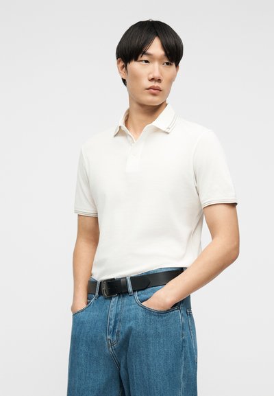 Νεαρός με κοντά μαύρα μαλλιά, φοράει λευκό πόλο πουκάμισο βιδωμένο σε μπλε τζιν με μαύρη ζώνη, τα χέρια στις τσέπες, κοιτάζοντας αριστερά.