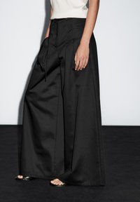 Black wide-leg trousers in a smooth fabric with a flat front, side pockets, and a drawstring waist. Folded hems and minimalist design.