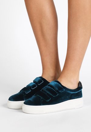 Stopy w ciemnoturkusowych aksamitnych trampkach na rzepy z trzema paskami i białą gumową podeszwą na białym tle.