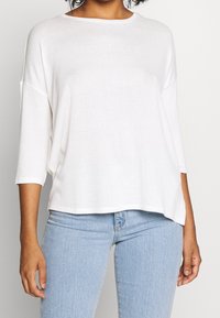 Kvinna i en lös, vit, långärmad topp och ljusblå högmidjade jeans som står mot en enfärgad bakgrund.