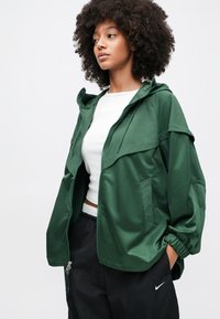 Νεαρή γυναίκα με σγουρά μαλλιά, φορώντας πράσινο μπουφάν με φερμουάρ, λευκό κοντό τοπ και μαύρο παντελόνι Nike, που στέκεται μπροστά σε απλό φόντο.