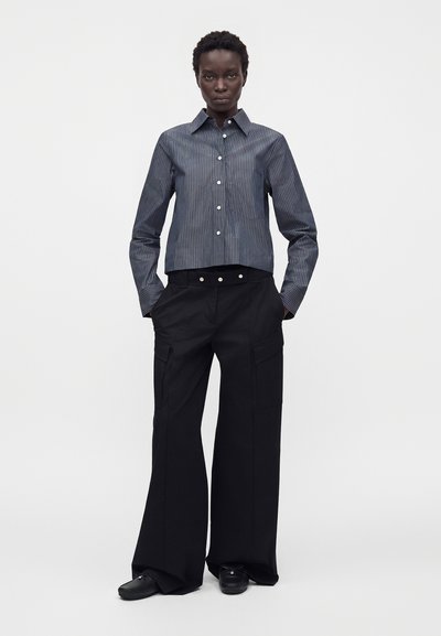 Femme portant une chemise rayée gris foncé boutonnée, un pantalon noir à jambes larges avec des poches cargo, et des chaussures noires, debout les mains dans les poches.
