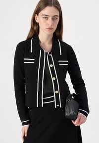 Black cardigan with white stripes along the collar and front, featuring gold buttons. Smooth texture, cropped design, layered over a black top.
