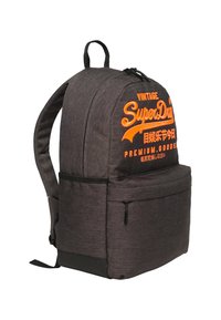 Mochila de tela negra con el logo naranja de "Superdry", bolsillo frontal con cremallera, correas acolchadas y fondo reforzado en material texturizado.