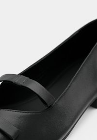 Μαύρο δερμάτινο παπούτσι φλατ με στρογγυλεμένη μύτη, με λουράκι στο πάνω μέρος και υφή φινιρίσματος. Χαμηλό τακούνι, μινιμαλιστικός σχεδιασμός.