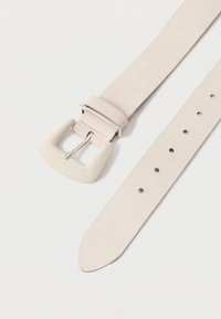 Ζώνη σε ανοιχτό μπεζ σουέντ με φαρδύ, καμπυλωτό κούμπωμα με ασημί φινίρισμα, ρυθμισμένη με πολλαπλές τρύπες κατά μήκος της ταινίας.