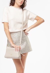 Beige leren crossbody tas met een gestructureerde afwerking en gouden hardware, vastgehouden door een persoon in een korte, glanzende zilveren rok en een lichte trui.