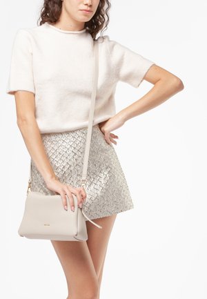 Borsa a tracolla in pelle beige con finitura strutturata e hardware color oro, tenuta da una persona in una gonna corta argentata lucida e un maglione chiaro.