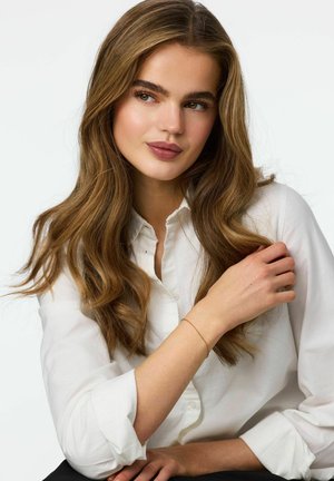 Modell med långt, vågigt brunt hår som bär en vit skjorta med knappar. Hon har ett tunt armband i guld runt handleden. Neutralt läppstift.