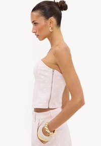 Femme en haut bustier à pois rose clair et pantalon, avec bracelets dorés et blancs et boucles d’oreilles créoles, cheveux en chignon, vue de profil.