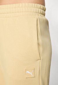 Beiga sweatpants med elastisk midja och sidofickor. Har en liten vit Puma-logotyp på en kvadratisk patch. Slät textur.