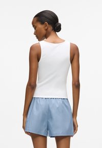 Baltas prigludęs marškinėliai be rankovių, derinami su šviesiai mėlynomis šortais, demonstruojantys lygią medžiagą, paprastą dizainą ir laisvą prigludimą.