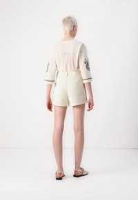 Top de lino beige claro con mangas de tres cuartos y acentos bordados, combinado con pantalones cortos beige de tiro alto con bolsillos traseros.