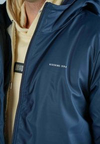 Veste imperméable bleu marine avec une texture lisse, dotée d'une capuche, d'une fermeture éclair et du logo imprimé 'REDSKINS 1984' sur la poitrine.