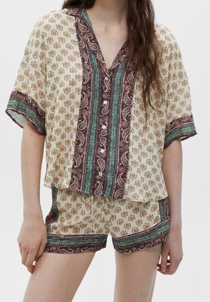 Kvinde iført en beige mønstret skjorte med knapper og matchende shorts med grønne, bordeaux og paisley kantdesign.