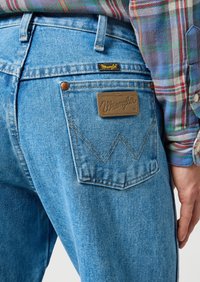 Lichtblauwe denim jeans hebben een bruine leren Wrangler-patch op de achterzak, met gele stiksels en een onderscheidend zigzagontwerp.