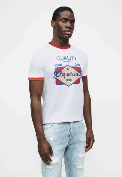 Νεαρός άνδρας που φοράει λευκό μπλουζάκι με κόκκινη επένδυση και μπλε γραφικό κείμενο, συνδυασμένο με ανοιχτό μπλε τζιν με σχισίματα, στέκεται μπροστά σε απλή φόντο.