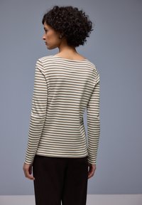 Frau mit lockigem Haar, die ein beige-weiß gestreiftes langärmliges Hemd und dunkle Hosen trägt, steht mit dem Rücken zum grauen Hintergrund.