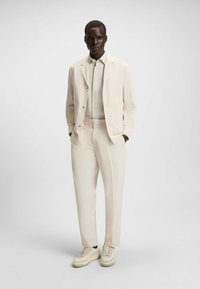 Completo color beige chiaro realizzato in tessuto morbido, con giacca su misura, pantaloni abbinati e sneakers crema con suola testurizzata.