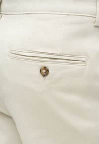 Κοντινό πλάνο σε μπεζ βαμβακερό παντελόνι με πίσω τσέπη τύπου welt και ένα καφέ κουμπί για κλείσιμο.