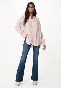 Camicia rosa chiaro a maniche lunghe con bottoni e tasca, abbinata a jeans a zampa d'elefante blu scuro e stivali alla caviglia beige, su uno sfondo bianco.