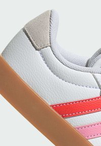 Zapatilla de deporte blanca de cuero con tres franjas horizontales en rojo, rosa y durazno. Talón de ante gris texturizado y suela de goma.