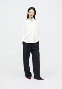 Camisa blanca de manga larga con botones, combinada con pantalones oscuros de pierna ancha y zapatos burdeos de punta; tejido suave, diseño minimalista.