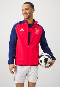 Chaqueta con cremallera en rojo y azul marino con un emblema de España, confeccionada en tejido ligero. Presenta el logo de Adidas y una textura suave. Pantalones cortos blancos y un balón de fútbol.