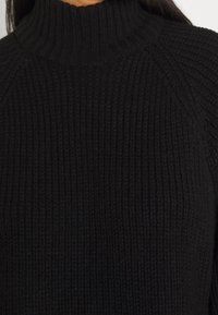 Pull noir en maille avec col montant, texture côtelée et manches raglan, mettant l'accent sur la chaleur et le confort dans le design.