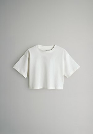 Balta īsām piedurknēm un īsā t-krekls ar apaļu dekoltē, kas izlikts uz vienkārša gaiši pelēka fona.