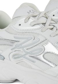 Zapatillas deportivas blancas con materiales de malla y cuero, que presentan acentos plateados y una suela texturizada. Los cordones en un tono neutro complementan el diseño.