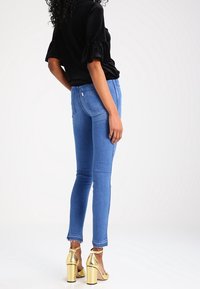 Jeans de denim azul skinny con dobladillo deshilachado y textura suave, combinados con tacones metálicos dorados con una correa. Top negro con mangas rizadas.