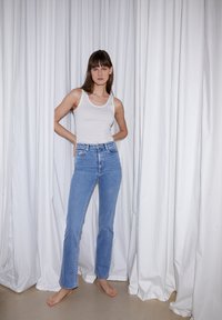 Top blanco sin mangas combinado con jeans de tiro alto en azul claro. La modelo está descalza apoyada contra una tela blanca caída. Diseño simple y sin adornos.
