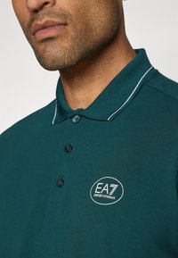 Temno zeleno polo majico iz teksturiranega blaga z dvema gumboma, belim obrobo okoli ovratnika ter logotipom EA7 na prsih.