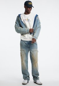 Giacca di jeans con accenti blu, felpa bianca con logo e pantaloni chiari strappati di colore blu. Sneakers bianche e grigie completano l'outfit.