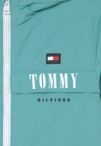 Teal jakke foran med hvid lynlås, Tommy Hilfiger logo patch med rød, hvid og blå flag, og fed hvid "TOMMY" tekst ovenfor broderet "HILFIGER" tekst.