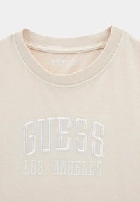 Beige organiskā kokvilnas T-krekls ar ribotu apkakli un izšūtu "GUESS LOS ANGELES" tekstu baltā krāsā. Mīksta tekstūra un vienkāršs dizains.