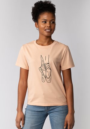 Jeune femme portant un T-shirt pêche avec un dessin au trait noir de chaussures de ballet pointes, associé à un jean bleu clair, debout devant un fond uni.