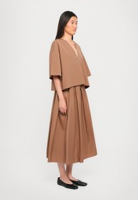 Brown cotton cropped top with wide sleeves and a V-neck, paired with a long, pleated brown skirt. Model wears black flat shoes.