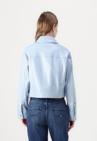 Giacca in pelle sintetica azzurro chiaro, tagliata, con colletto a punta, polsini con bottoni e una texture liscia e lucida. Abbinata a jeans in denim scuro.