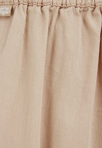 Beige tyg med en strukturerad yta och detaljerad midjeresår. Materialet verkar mjukt med subtila veck och en etikettaccent.