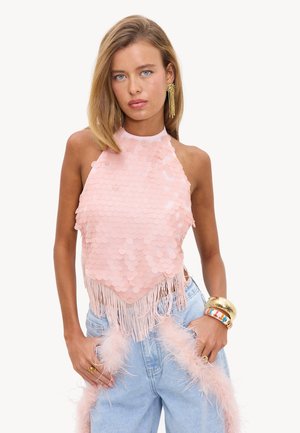 Jeune femme aux cheveux blonds raides porte un top dos-nu à sequins rose pâle avec des franges et un jean bleu clair, accessoirisé avec des boucles d'oreilles et bracelets dorés.