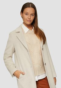 Beige Wollmantel mit Reverskragen und zwei Fronttaschen, über einem cremefarbenen Strickpullover getragen. Das Model hat langes, glattes Haar.