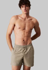 Pantaloni da bagno beige con vita elasticizzata e dettaglio con cordoncino, realizzati in tessuto leggero, dotati di tasche laterali e texture liscia.