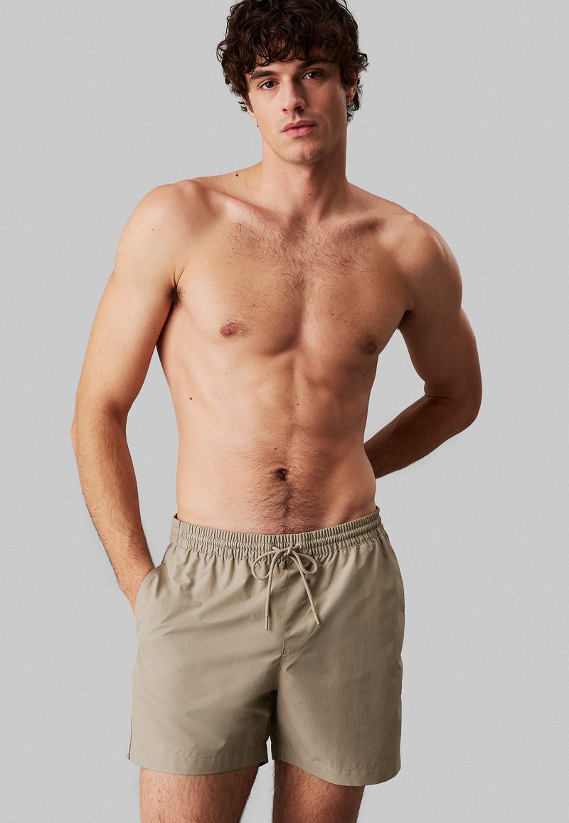 Pantaloni da bagno beige con vita elasticizzata e dettaglio con cordoncino, realizzati in tessuto leggero, dotati di tasche laterali e texture liscia.