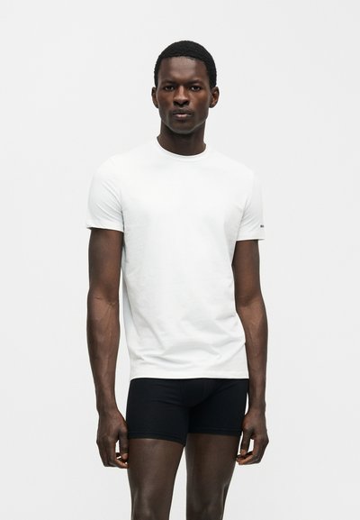 Νέος άνδρας στέκεται όρθιος φορώντας ένα απλό άσπρο κοντομάνικο μπλουζάκι και στενά μαύρα σορτς πάνω σε λευκό φόντο.