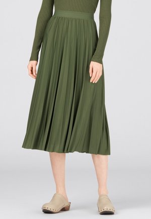 Vrouw draagt een olijfgroene geplooide midirok, een geribd topje met lange mouwen en beige gespikkelde klompen, staand tegen een grijze achtergrond.
