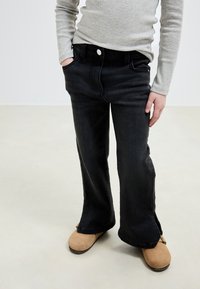 Schwarze Schlagjeans mit klassischem Fünf-Taschen-Design, versehen mit einem Knopf- und Reißverschluss, kombiniert mit einem hellgrauen Langarmshirt.