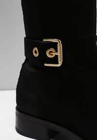 Bottine en daim noir avec boucle dorée et rivet doré estampillé sur le côté au-dessus du talon empilé.